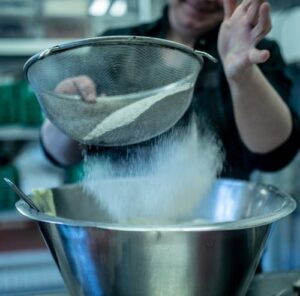 A female baker sifting flour into a metal bowl.